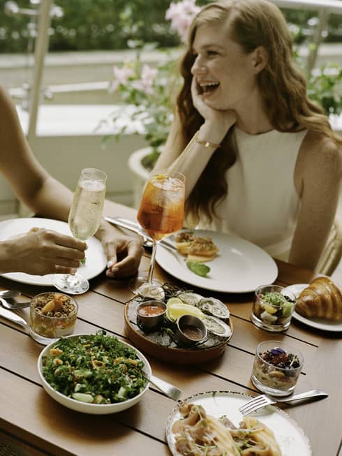 Laughing guests enjoying cold beverages, raw oysters with sauces, smoked salmon, a kale salad, muesli and croissants.