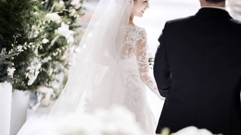 A bride in a white, lace, long sleeve gown smiles as she walks down the aisle with her groom. They are surrounded by artfully arranged white flowers.