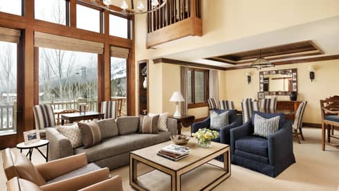 Spacious resort living area with a grey sofa, navy armchairs and large windows offering snowy mountain views, connected to a dining area with striped chairs