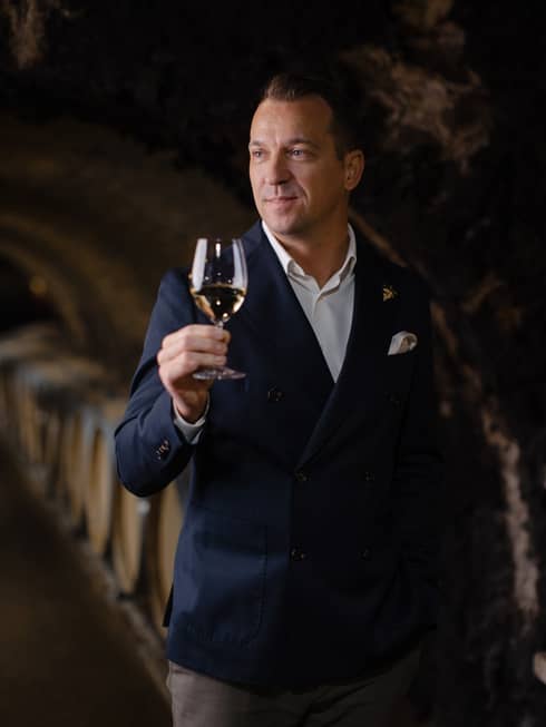 Person in a dark suit holding a glass of white wine in a wine cellar