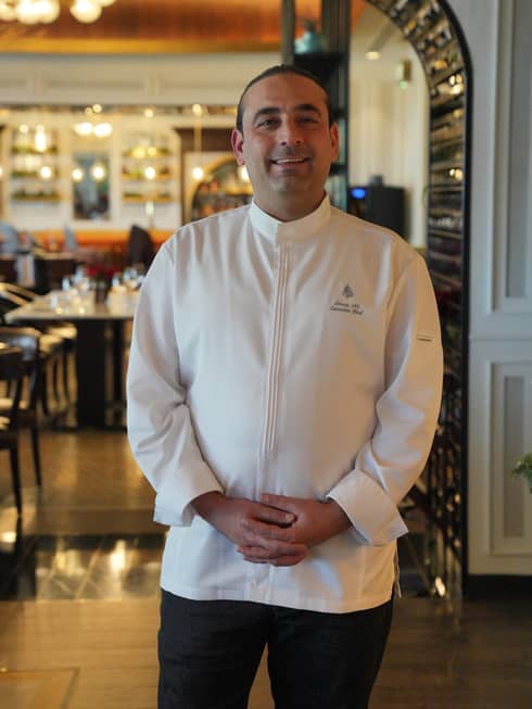 Chef wearing a white chef's jacket and black pants stands in a resaturant dining room smiling