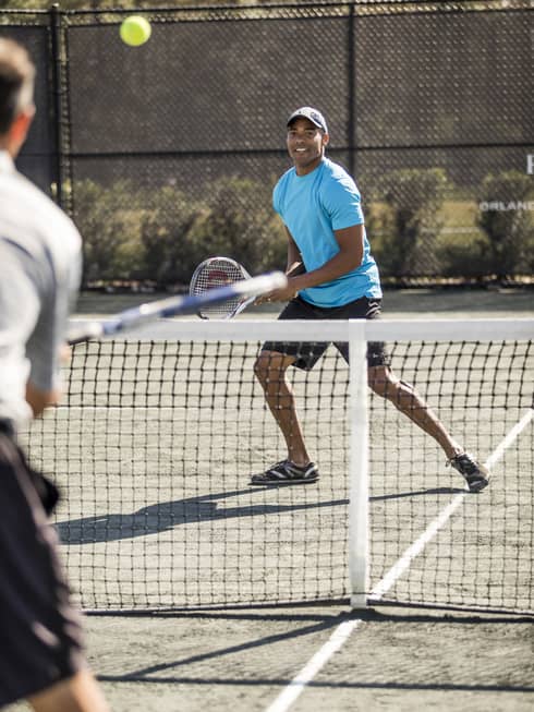 Man wearing blue T-shirt and hat holds racket as tennis ball flies over net