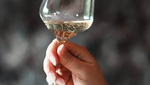 Close-up of a hand holding a tilted glass of white wine against a dark, blurred background.