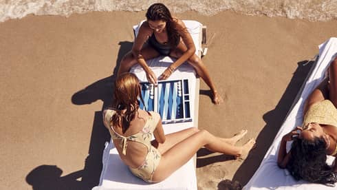 People relaxing on lounge chairs at a sandy beach, with two of them playing a backgammon game