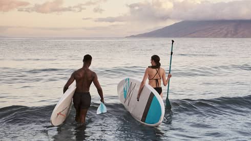Two people wading into the ocean with paddleboards during a calm sunset