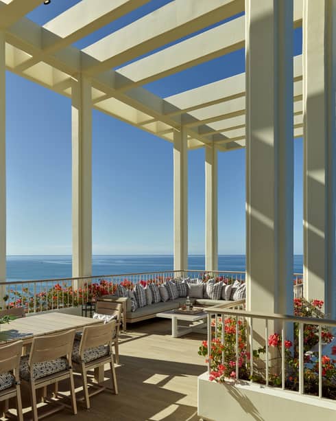 A spacious terrace with a pergola overlooking the ocean, featuring a large dining table, comfortable seating and vibrant flower arrangements under a clear blue sky