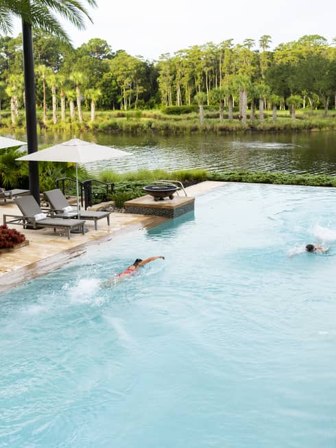 Two people swim in a large pool with two sets of steps leading into it. The pool is flanked by deck areas with lounge chairs, umbrellas, and palm trees and other plantings. A natural-looking body of water and other plants and trees are in the background.