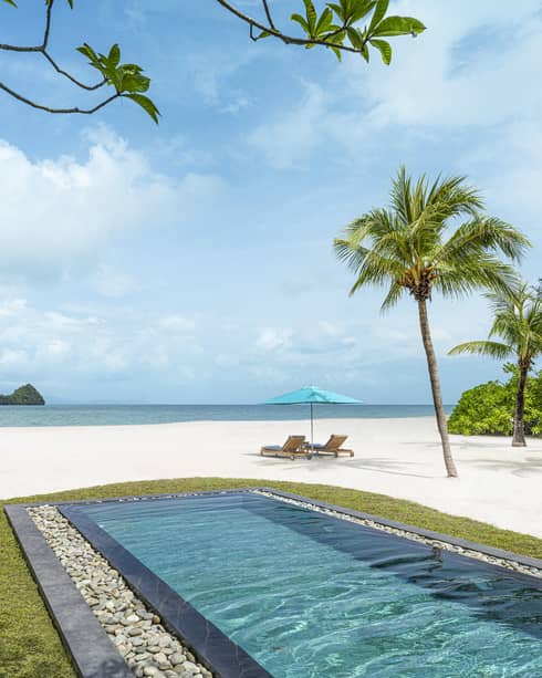Rectangular in-ground pool jutting out to white-sand beach dotted with palm trees with two lounge chairs and umbrella along blue waters