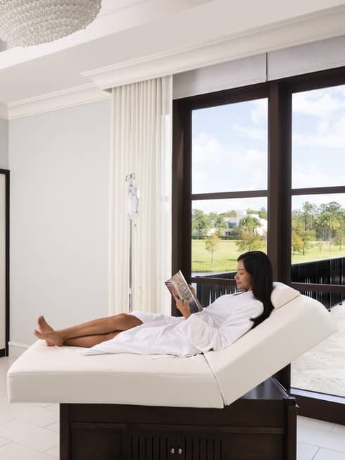 Woman wearing white bath robe reads magazine in spa lounge chair