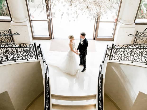 Aerial view of a bride and groom standing in the middle of a grand staircase below an ornate crystal chandelier