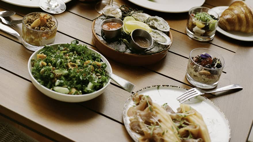 Laughing guests enjoying cold beverages, raw oysters with sauces, smoked salmon, a kale salad, muesli and croissants.