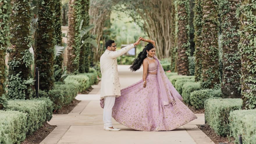A groom wearing all white twirls his bride wearing a lavendar Indian wedding dress on a walkway lined with green hedges and towering palm trees creating a canopy of green above