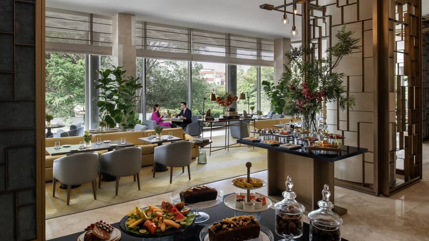 Two people sit a small table by window in room with metal d�cor and various types of food on table display