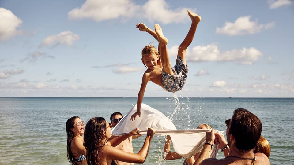 A group enjoys a sunny beach day, tossing a child into the air with a white sheet. The child smiles mid-air against clear skies.