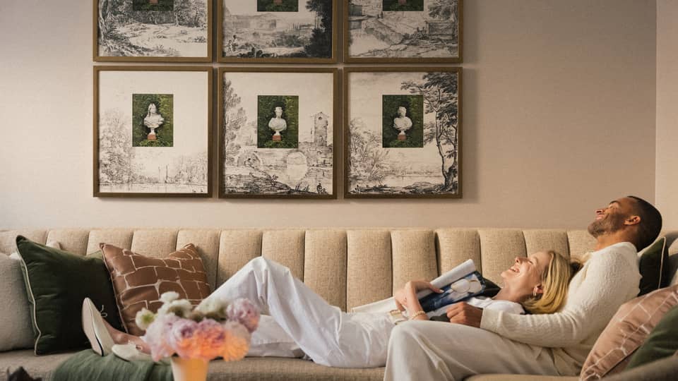 Two guests laying on the couch in a guest room with picture frames on the wall and a bouquet of flowers on the coffee table