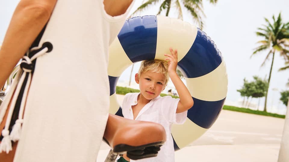 Young child wearing green swim trunks and a white shirt holds a blue-and-cream pool float while walking next to adult wearing a cream-coloured beach cover up and walking next to a bicycle