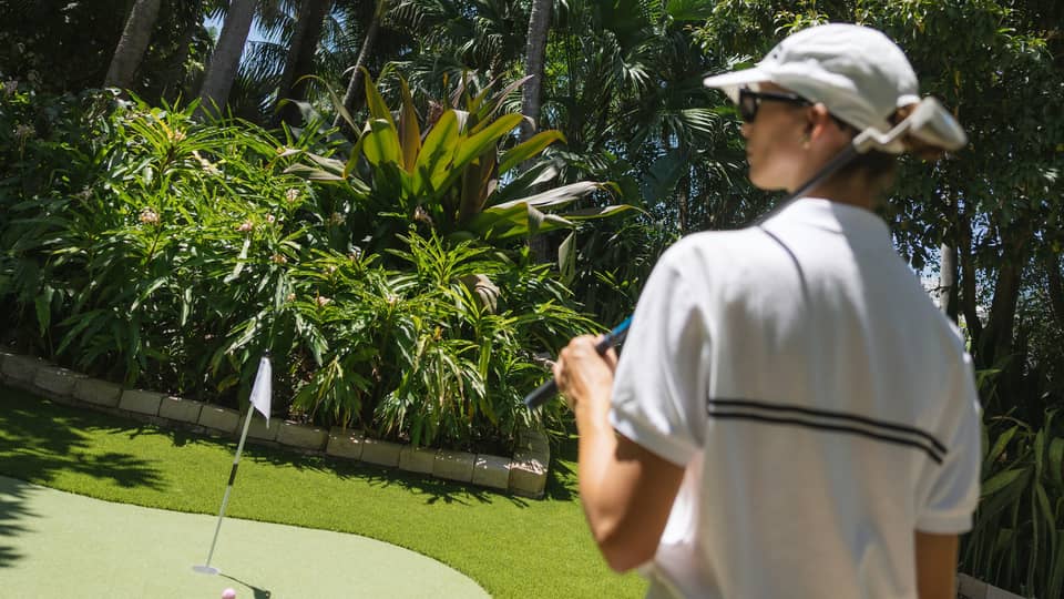 Golfer wearing white sweater vest, white skirt and white hat walks toward a putting green surrounded by palm trees