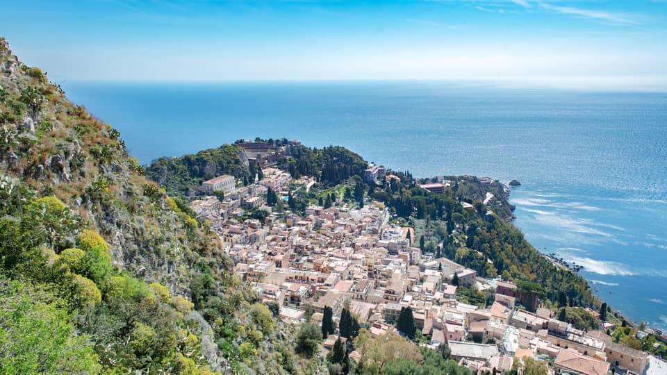 Aerial view of a coastal town with clustered buildings, lush greenery and the blue expanse of the ocean in the background.