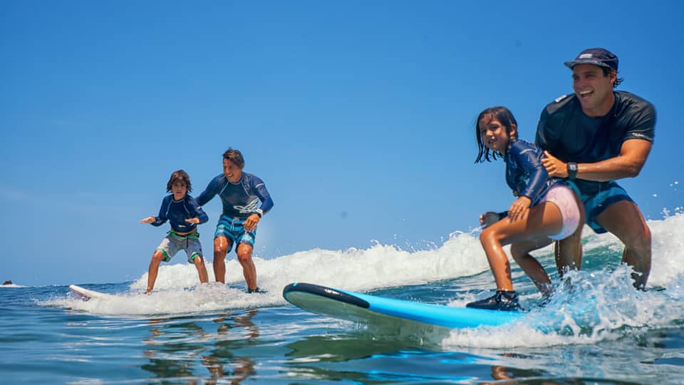 A pair of smiling adults and children standing on two surfboards, wearing swimsuits and rash guards, riding a small wave.