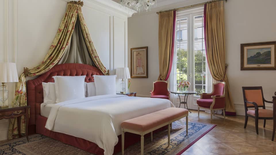 Lavish hotel suite bedroom, featuring a king bed with draped canopy and red armchairs by a window