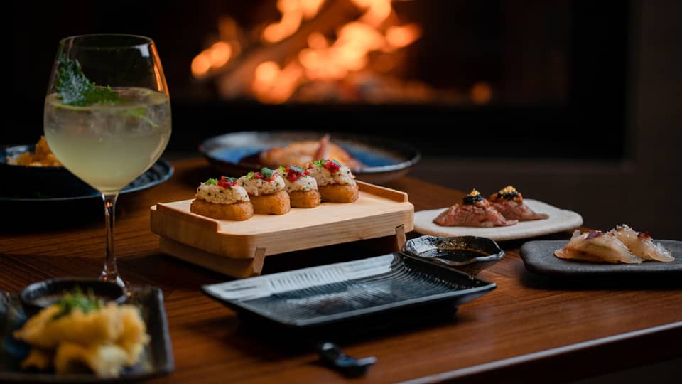 A cocktail and appetisers placed on a wooden table with a fireplace glowing in the background, creating a cosy dining atmosphere