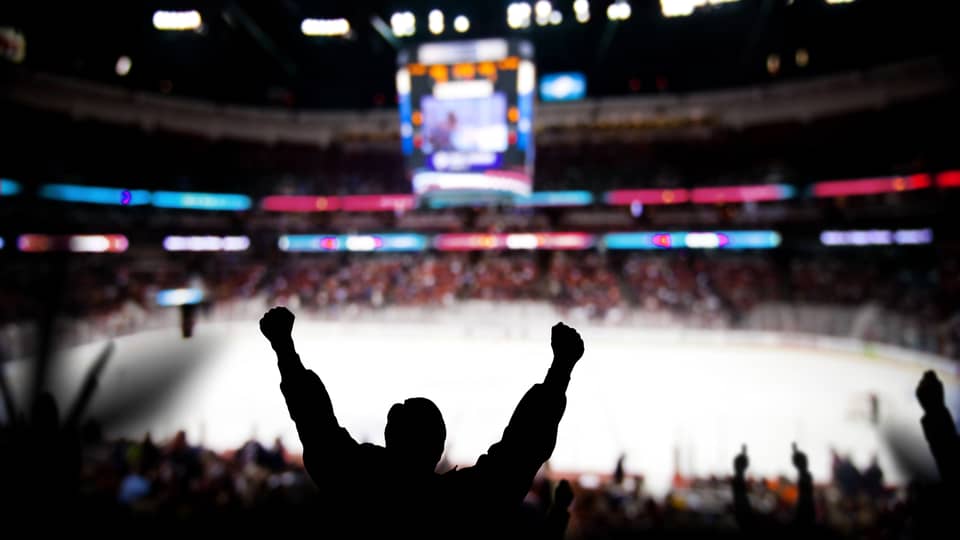 Silhouette of a fan cheering with raised arms at a hockey game in a crowded arena, with the ice rink and scoreboard in the background.