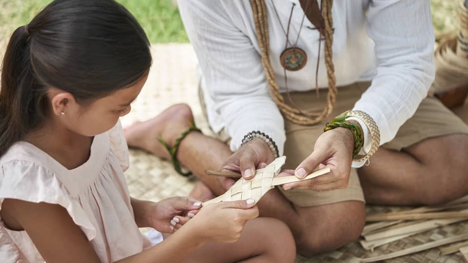 An adult shows a young girl how to weave a basket