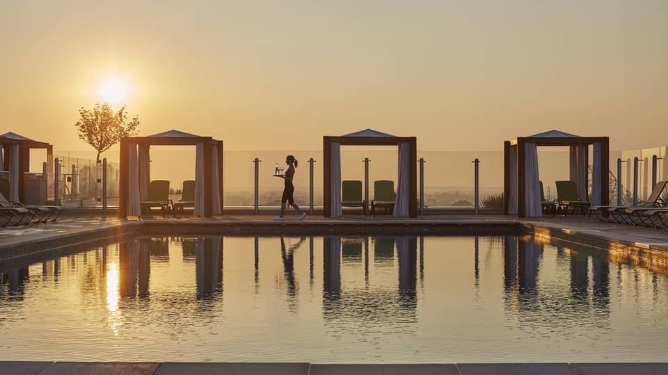 A server walking by a rooftop pool with cabanas at sunset and carrying drinks, with the golden sky reflecting on the water in a luxury hotel terrace setting