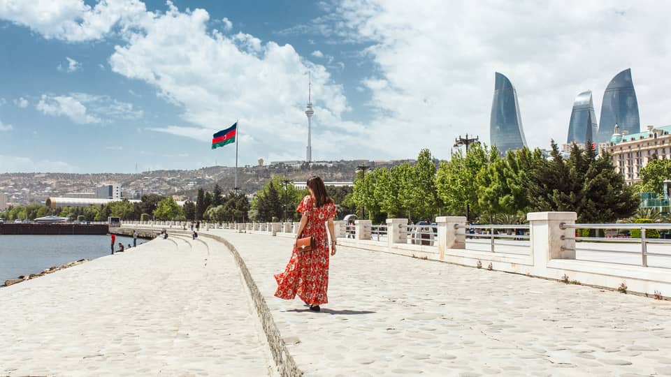 Woman wearing red dress walks along stone waterfront, skyscrapers in background