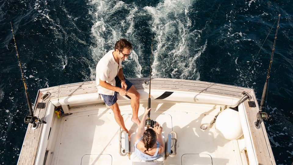 Two people sit at the back of a fishing boat, with one person holding a fishing rod and the other sitting close by on the edge of the boat