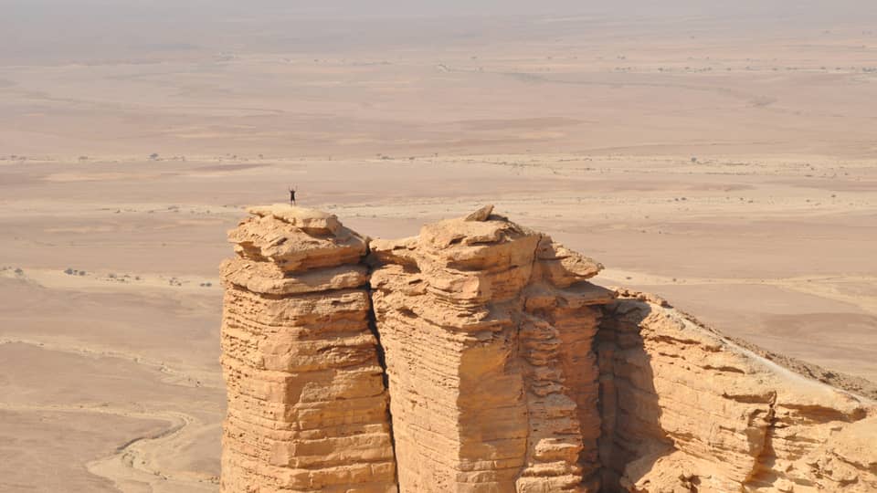 An aerial view of a giant golden cliff surrounded by a dry desert and a small silhouette of a person standing on top of it.