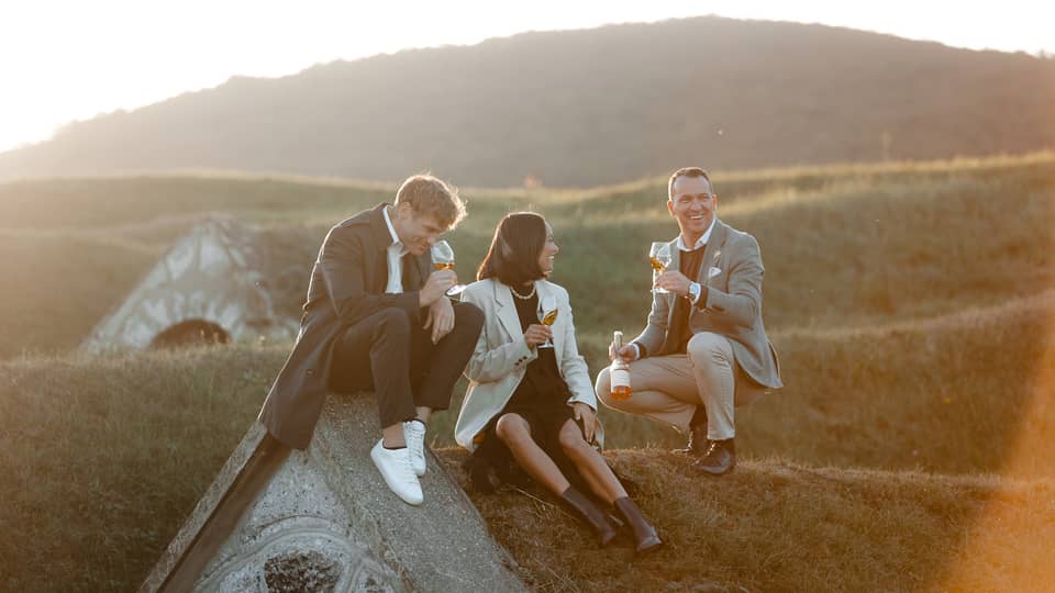 A group of people sitting outdoors on a grassy hill, enjoying glasses of wine together in the warm evening light.