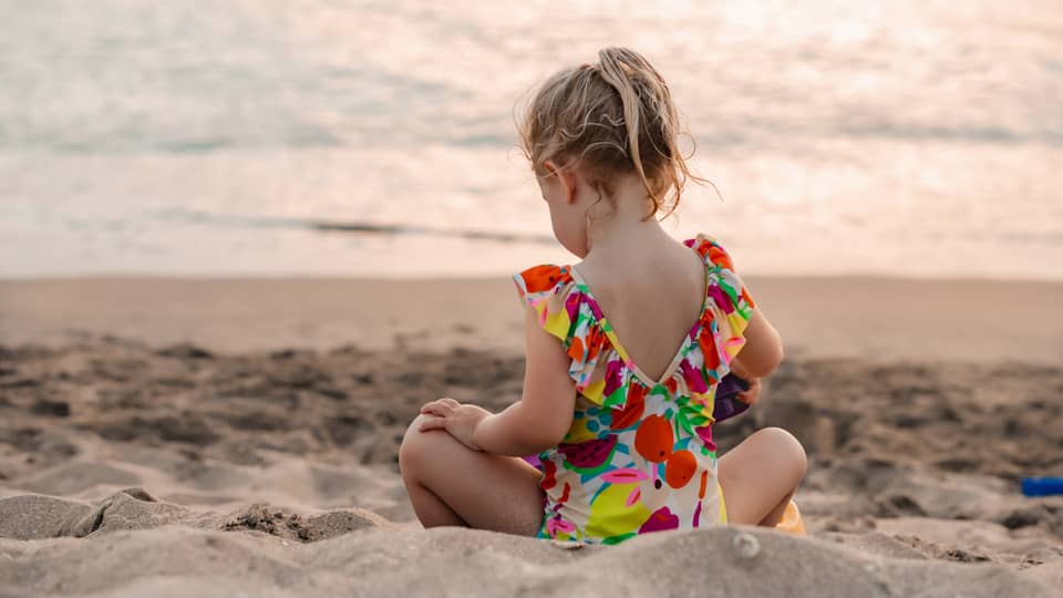 Young child sits on a sandy beach facing the water