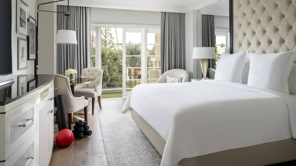 A hotel room with large open window, bed with white sheets, dresser, two chairs, an exercise ball and weights.