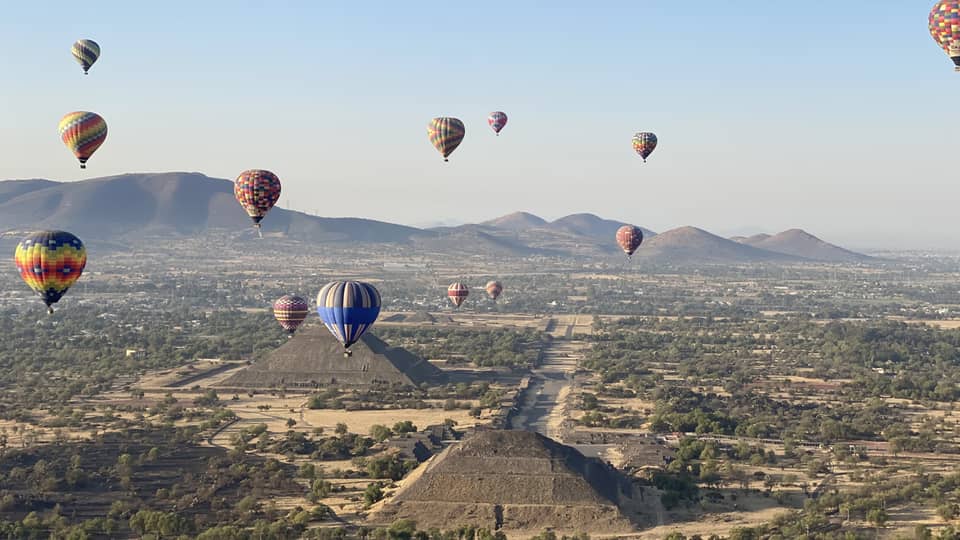 Hot-air balloons floating over old temples in a dessert covered in shrubs.
