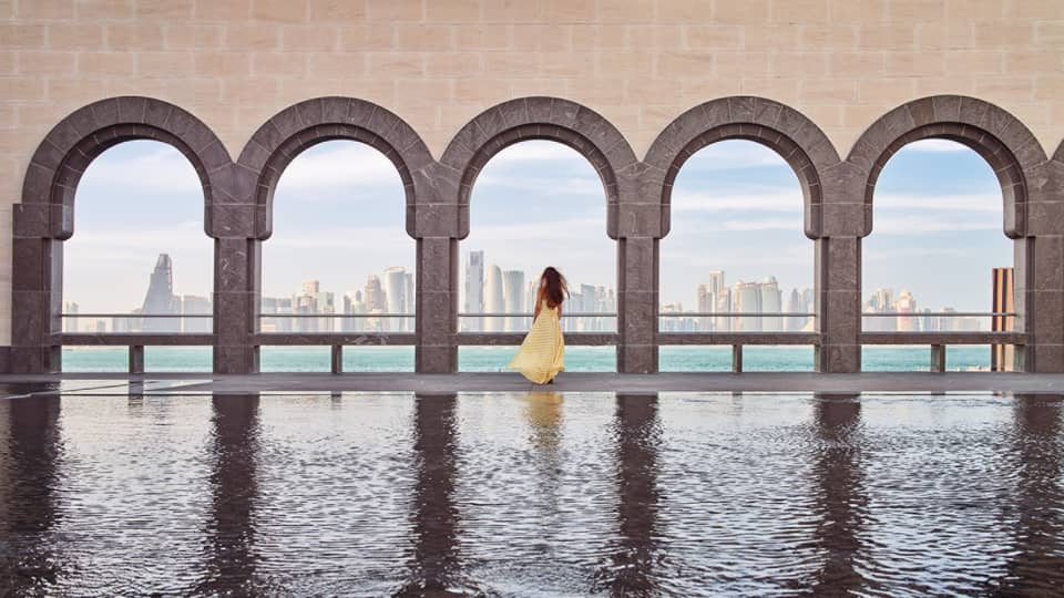 Woman wearing a long flowing yellow dress stands at the edge of a fountain beneath a wall of archways looking out onto the sea and a city skyline