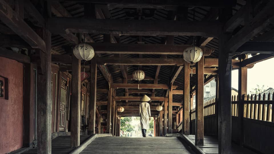 A person wearing traditional Vietnamese attire and a conical hat walks through an ancient wooden outdoor hallway decorated with lanterns.