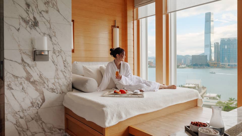 Woman in spa bathrobe sits on windowside chaise looking out to Victoria Harbour at Four Seasons Hotel Hong Kong