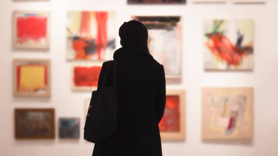 Silhouette of back of woman with hat bag looking at gallery wall with colourful art