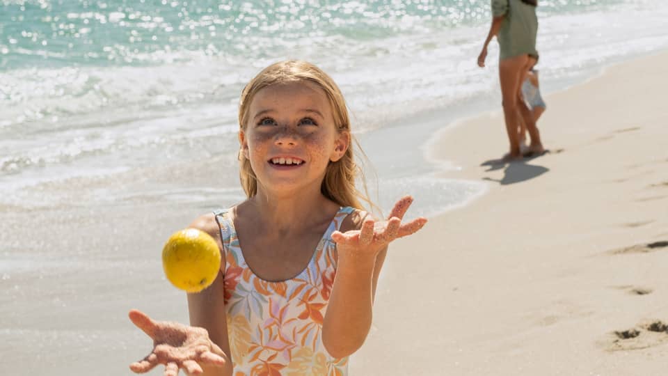 Child juggling lemons on a sunny beach with gentle ocean waves in the background, while another person walks along the shoreline in the distance.