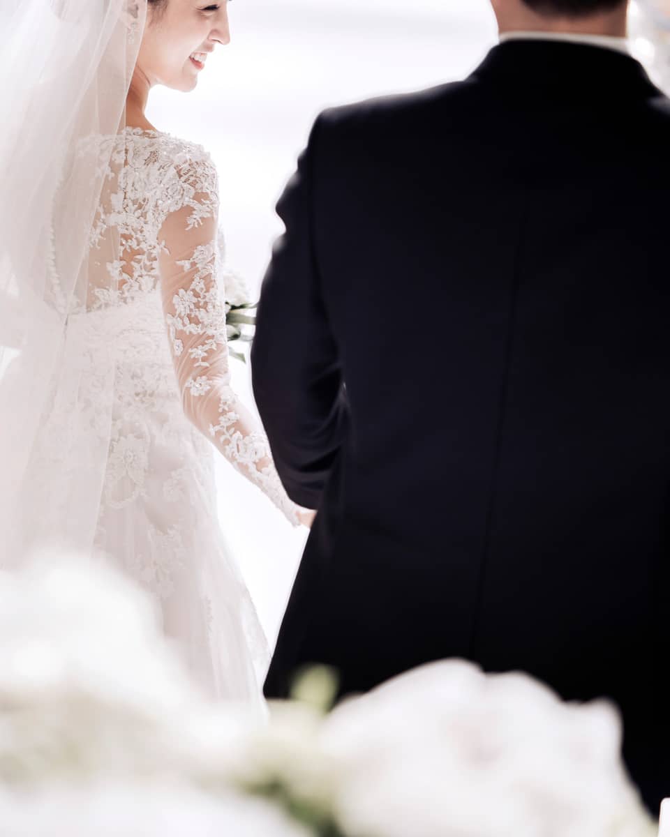 A bride in a white, lace, long sleeve gown smiles as she walks down the aisle with her groom. They are surrounded by artfully arranged white flowers.