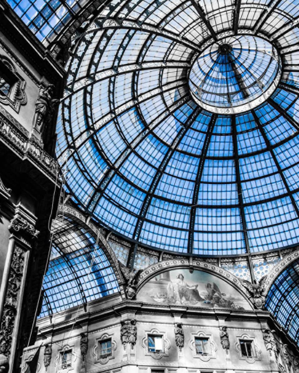 Interior view of the Galleria Vittorio Emanuele II in Milan, showcasing its grand glass dome ceiling and ornate architectural details in a mix of black and white with blue-tinted glass.