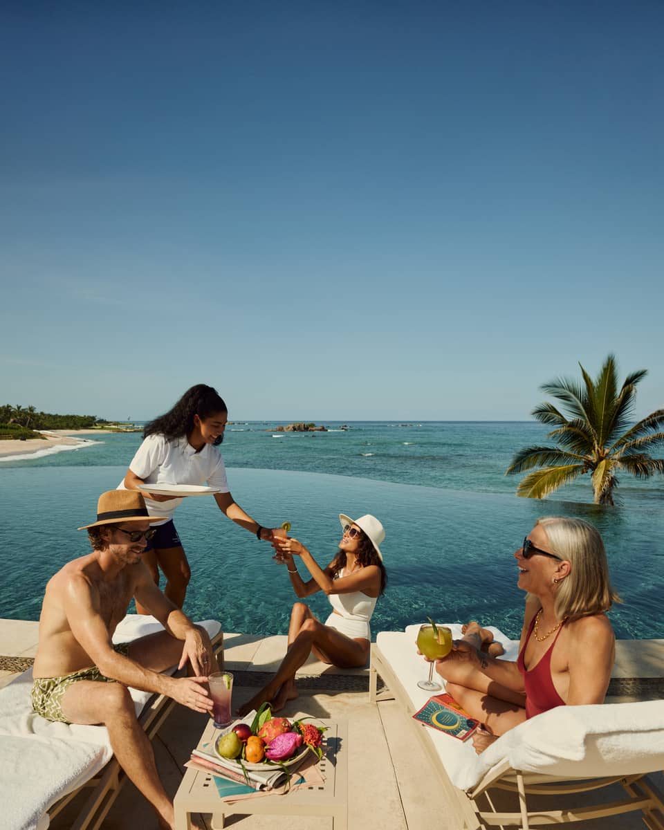 Three people mingle over cocktails next to an ocean-view resort pool, as server serves a drink