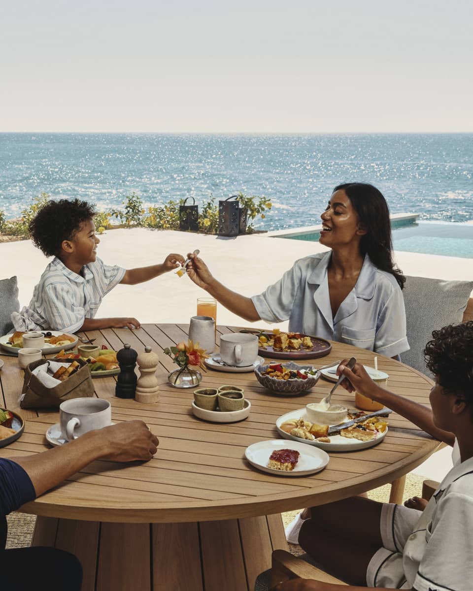 Family enjoying breakfast together at a round wooden table on an ocean-view terrace at a luxury resort