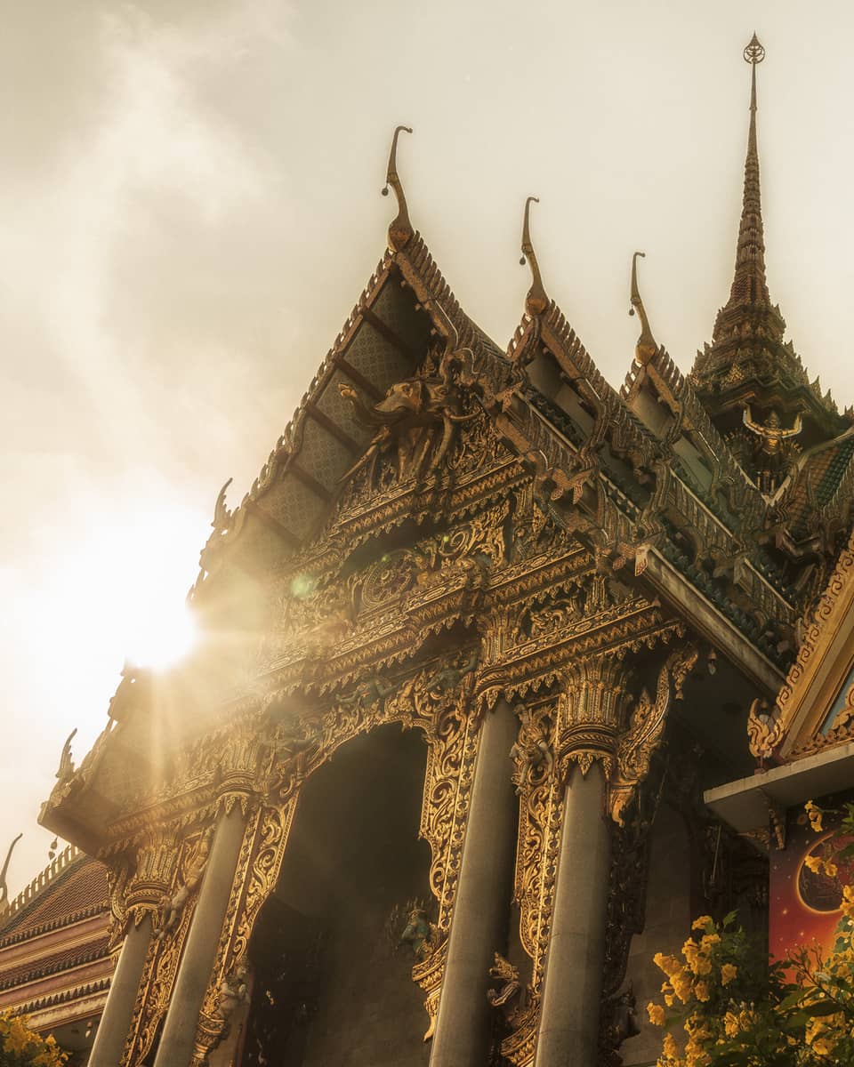 Intricately designed temple with ornate carvings and tiered roofs, illuminated by sunlight, with a tree and yellow flowers in the foreground