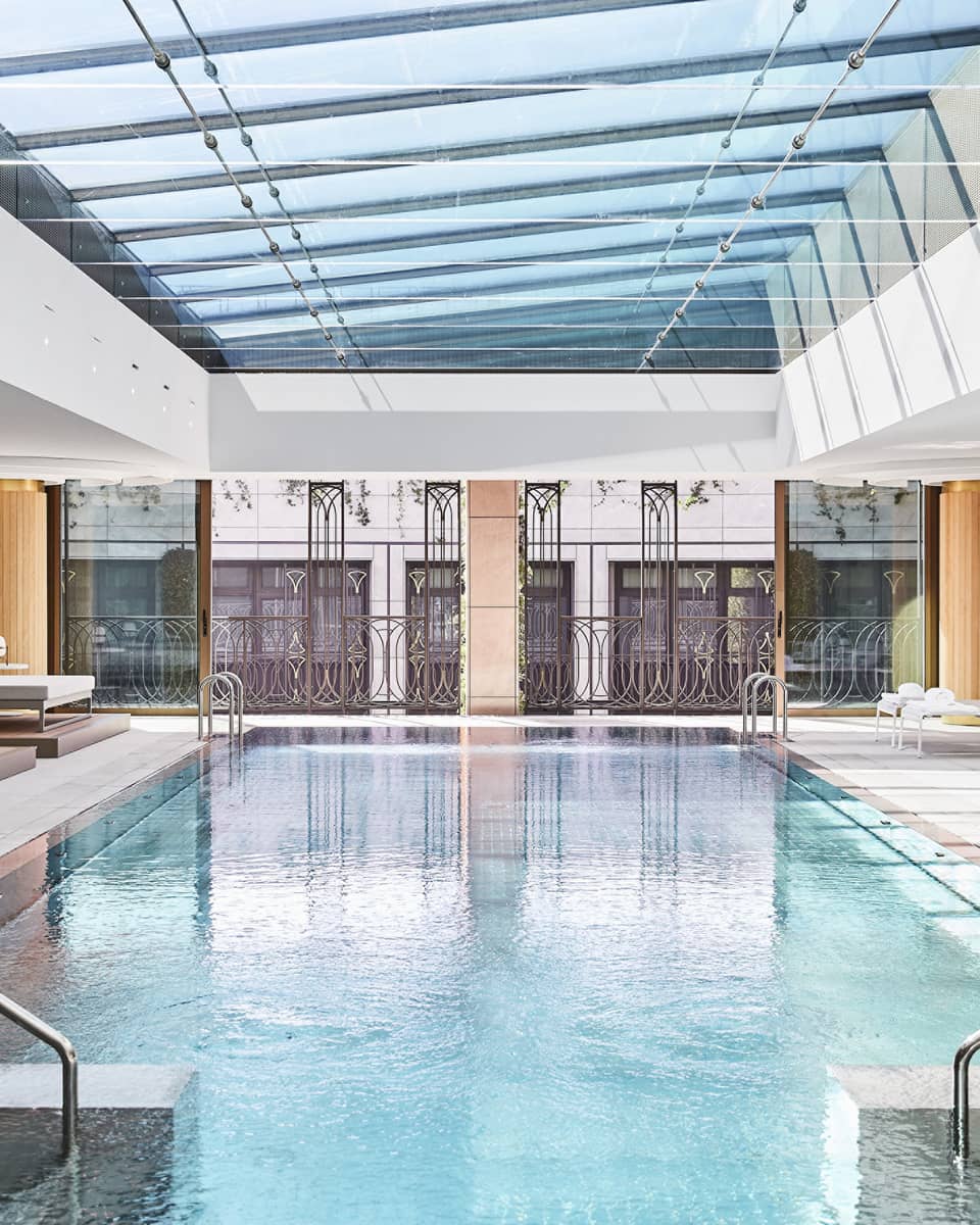 Luxury hotel indoor pool area with glass ceiling, metal railing accents and lounge chairs, surrounded by large windows and architectural detailing