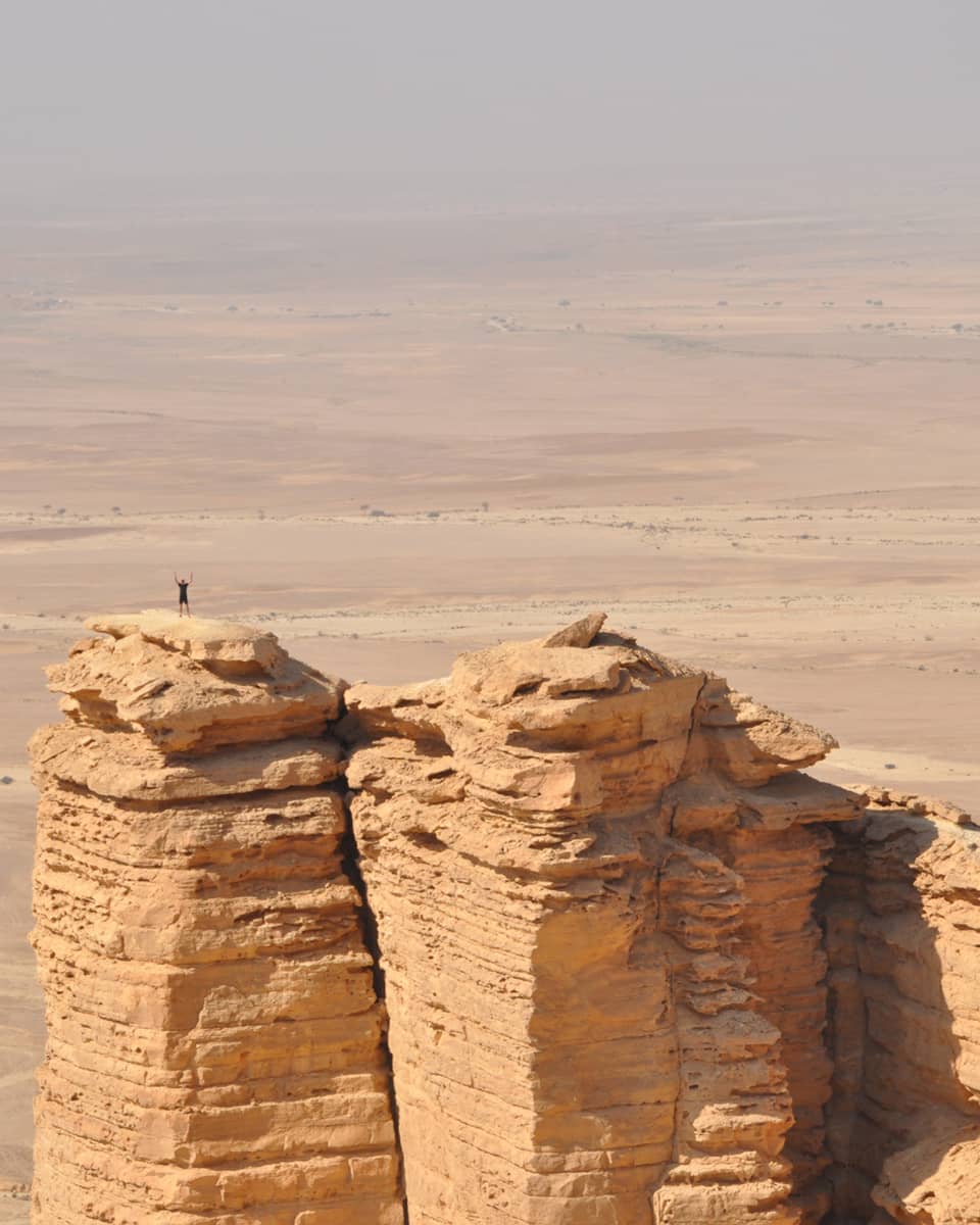 An aerial view of a giant golden cliff surrounded by a dry desert and a small silhouette of a person standing on top of it.
