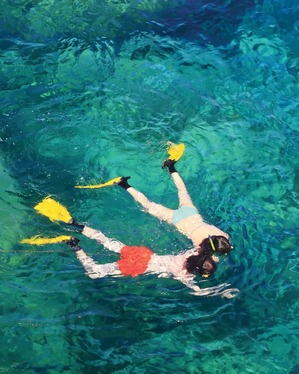 View from above of two guests in flippers snorkelling hand in hand, surrounded by crystal turquoise waters.