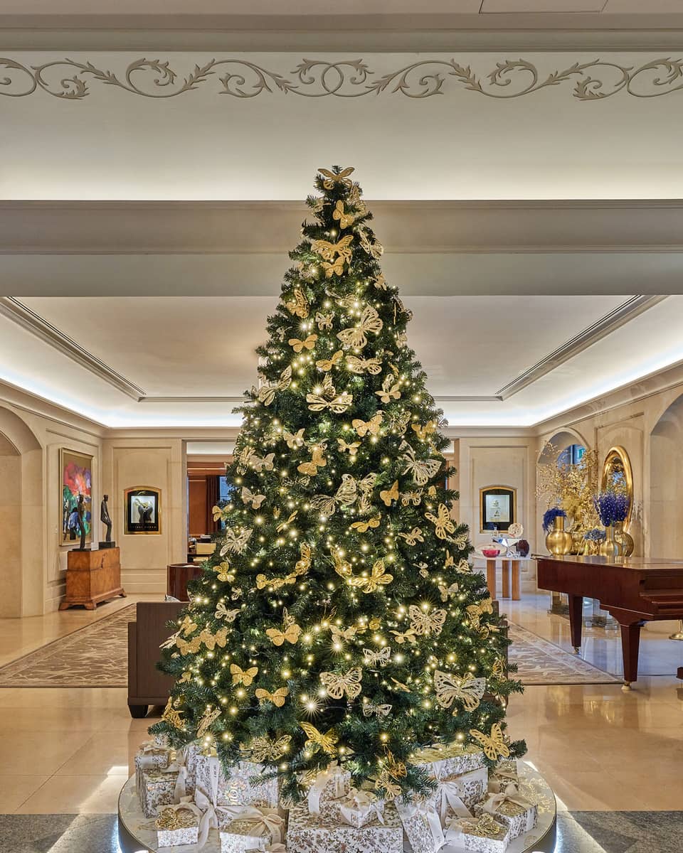 Luxurious hotel lobby, featuring a large elegantly decorated Christmas tree with gold ornaments and lights, surrounded by Christian Dior gift boxes and modern artwork on the walls.