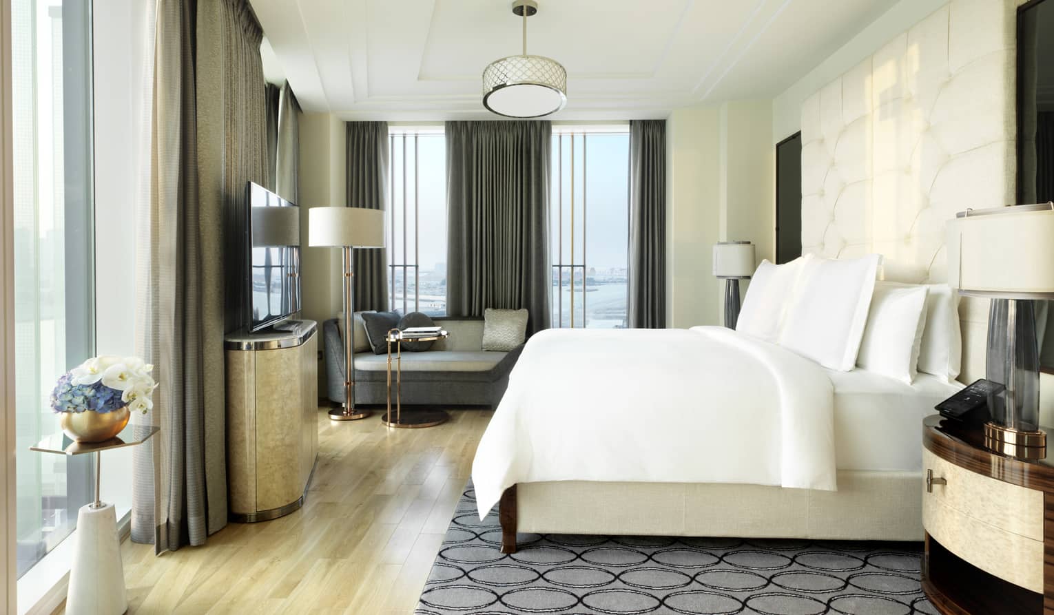 Luxurious hotel bedroom with a white bed, tufted headboard, a cozy sitting area, large windows with city and water views and elegant decor accents